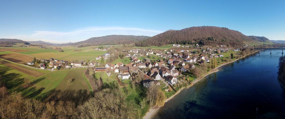 A riverside village with houses and fields, a bridge, and hills in the background.
