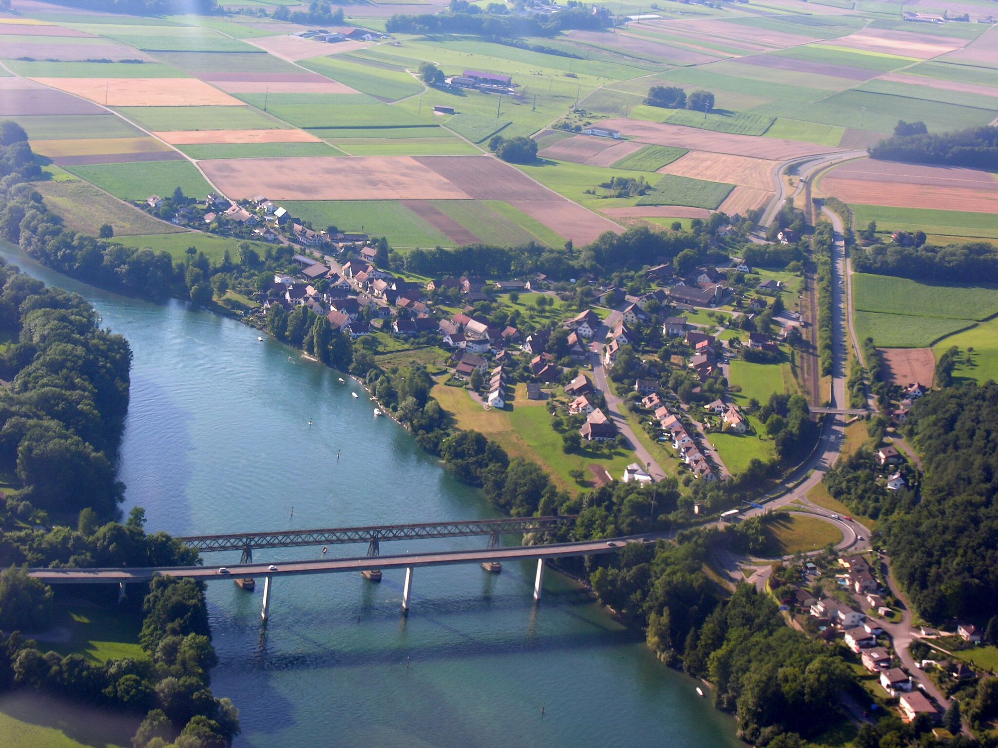 An aerial view of a river town with a bridge, surrounded by fields and greenery.
