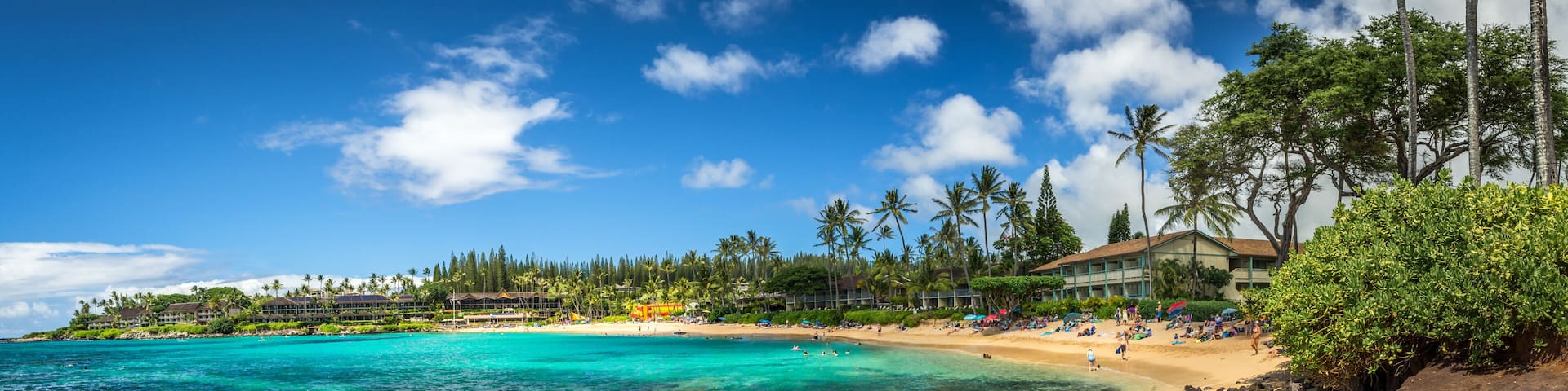 A tropical beach with clear turquoise waters, a rocky shoreline, and a resort with palm trees in the background.