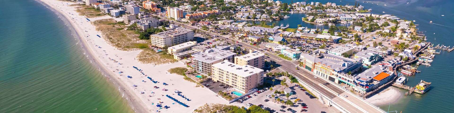 An aerial view of a coastal city with a long sandy beach, a marina, and a bridge connecting the mainland to an island.