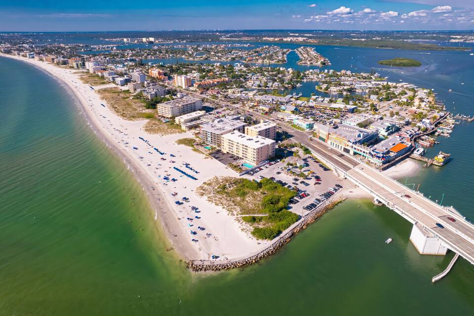 Eine Luftaufnahme einer Küstenstadt mit einem langen Sandstrand, einem Yachthafen und einer Brücke, die das Festland mit einer Insel verbindet.