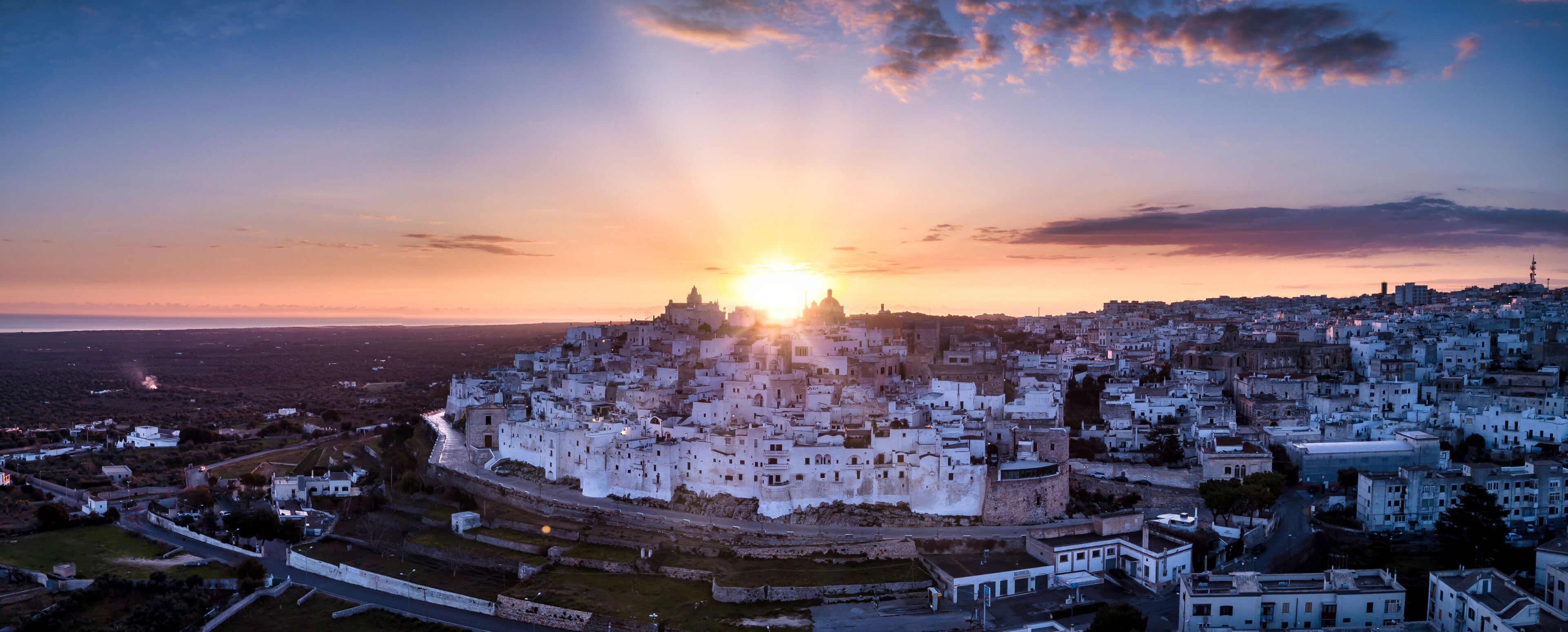 A cityscape with a prominent hilltop fortress, surrounded by densely packed buildings, during sunset.