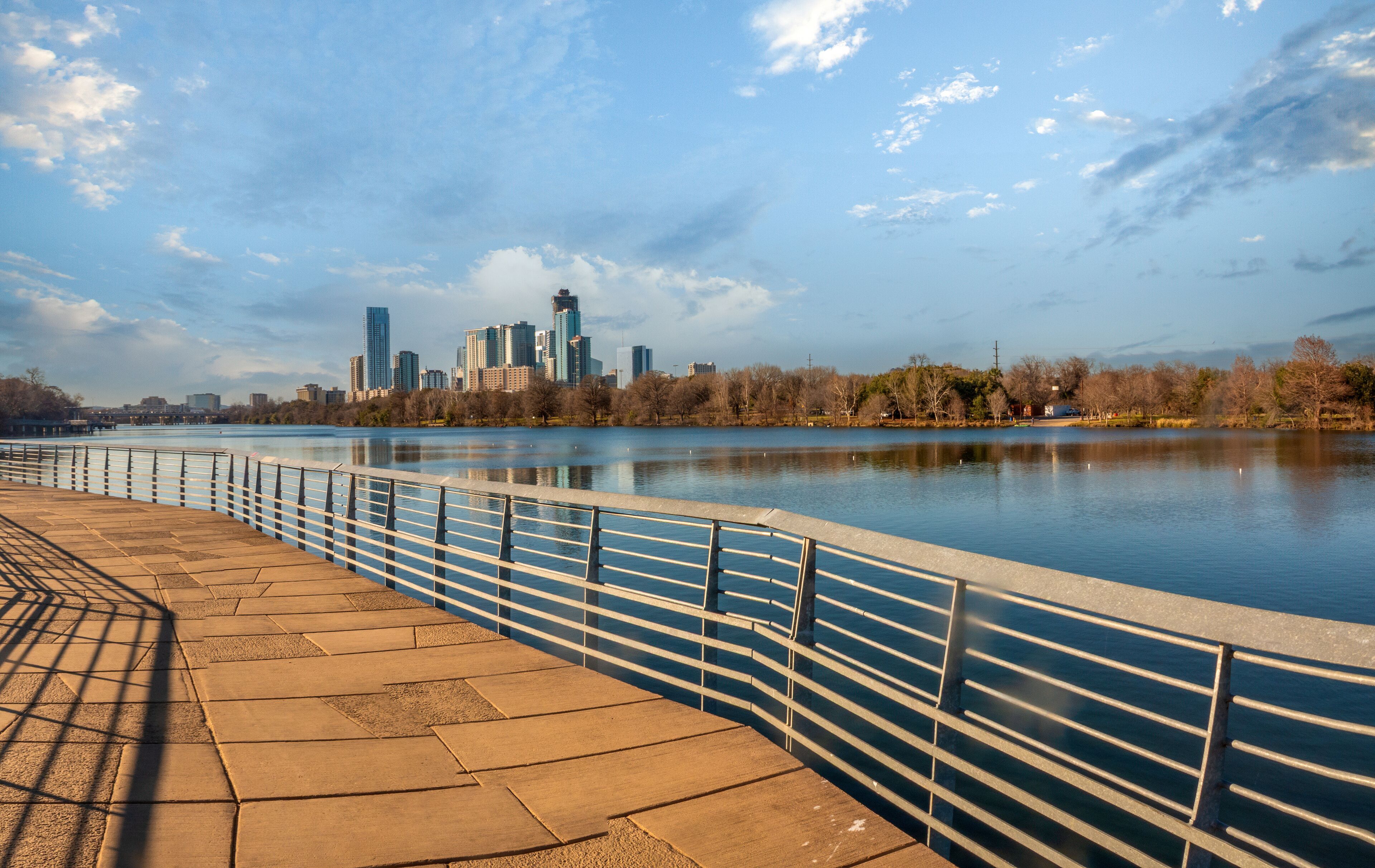 Un paseo ribereño con barandilla de metal, un río tranquilo y el perfil urbano de una ciudad al fondo.