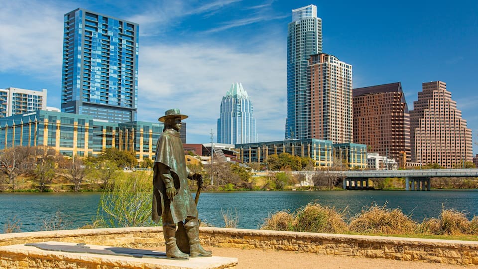 A statue of a man in a hat and boots by a river with a cityscape in the background.