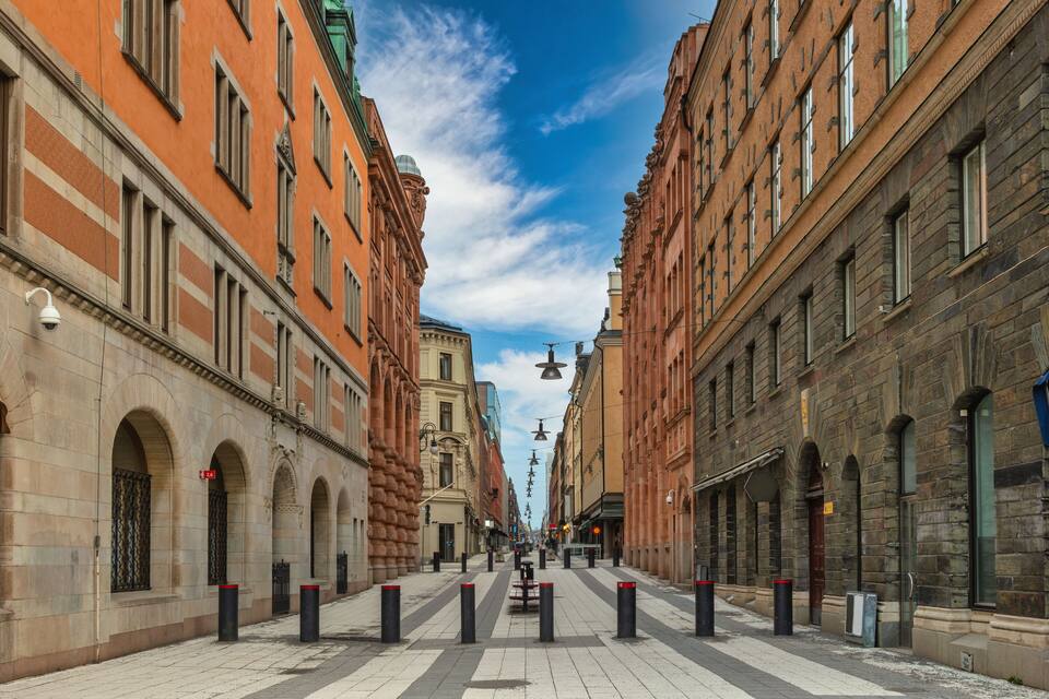 A wide, historic street lined with red and beige buildings, featuring arched windows and a clear blue sky.
