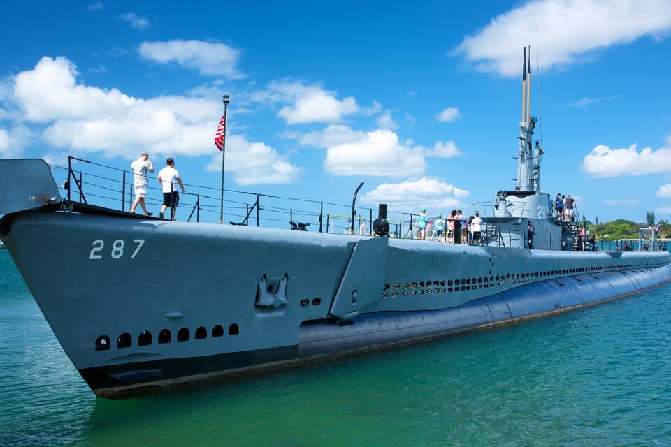 Un submarino con el número 287, atracado en un muelle con gente a bordo y un cielo azul despejado al fondo.