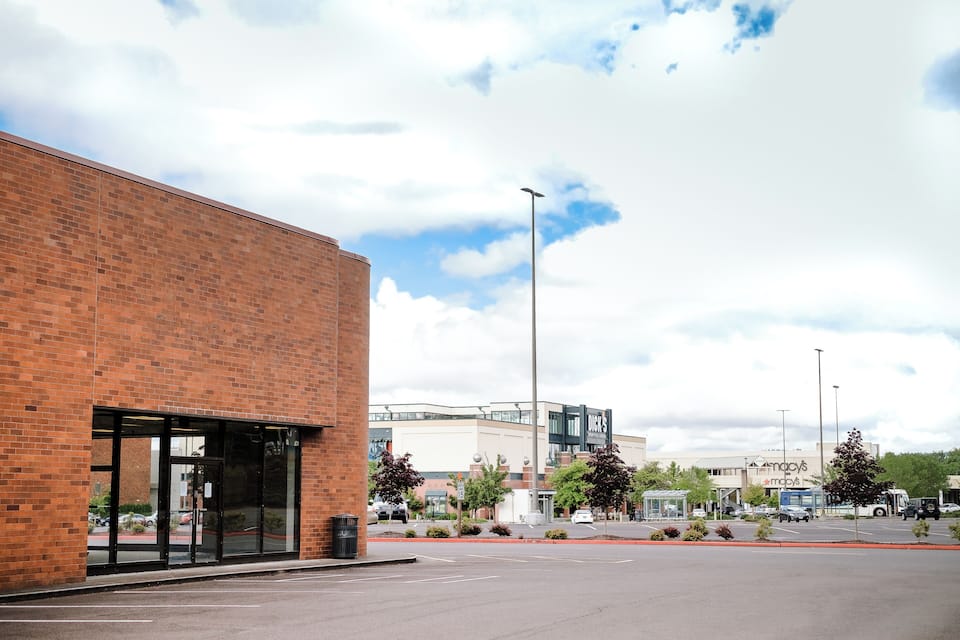 A parking lot with a brick building, a bus stop, and a shopping mall in the background.