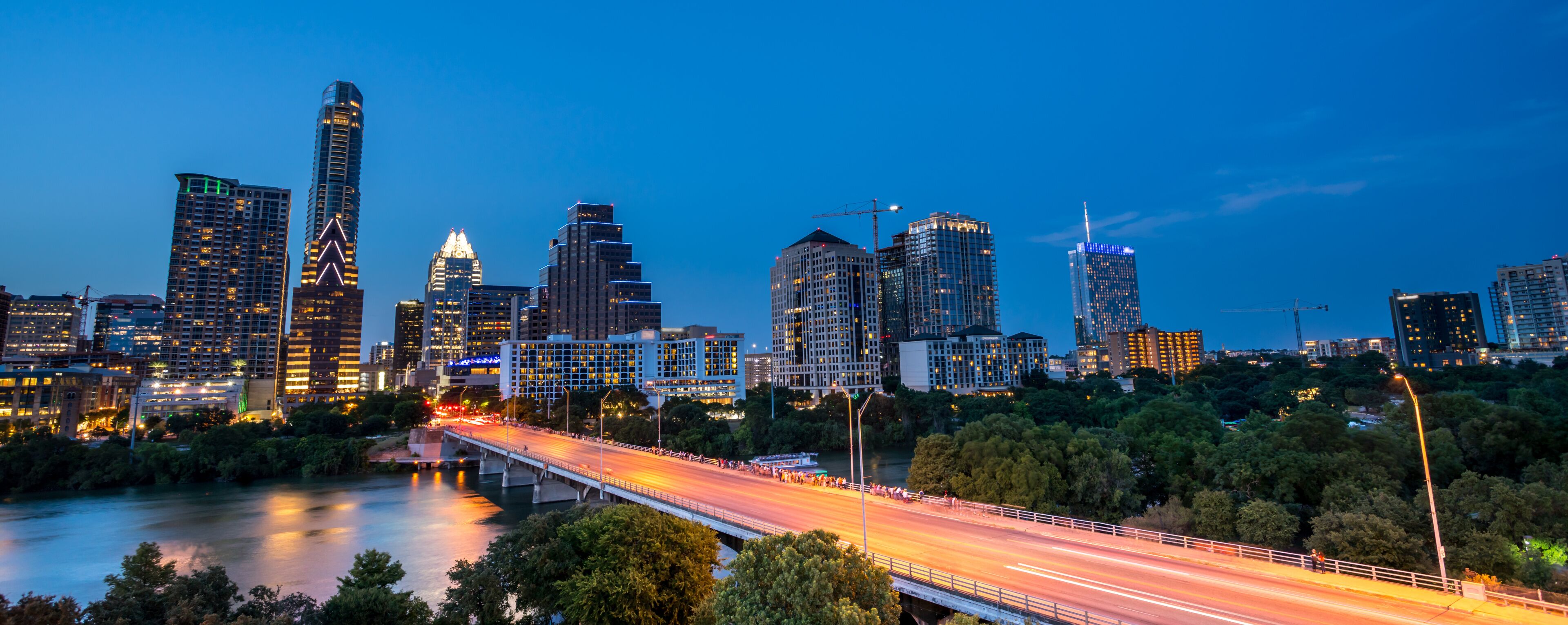 A city skyline at dusk with illuminated buildings and a bridge over a river.