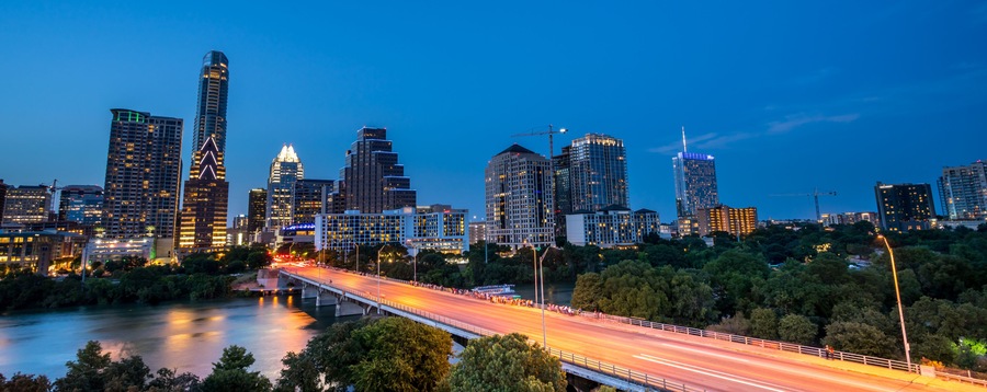 A city skyline at dusk with illuminated buildings and a bridge over a river.