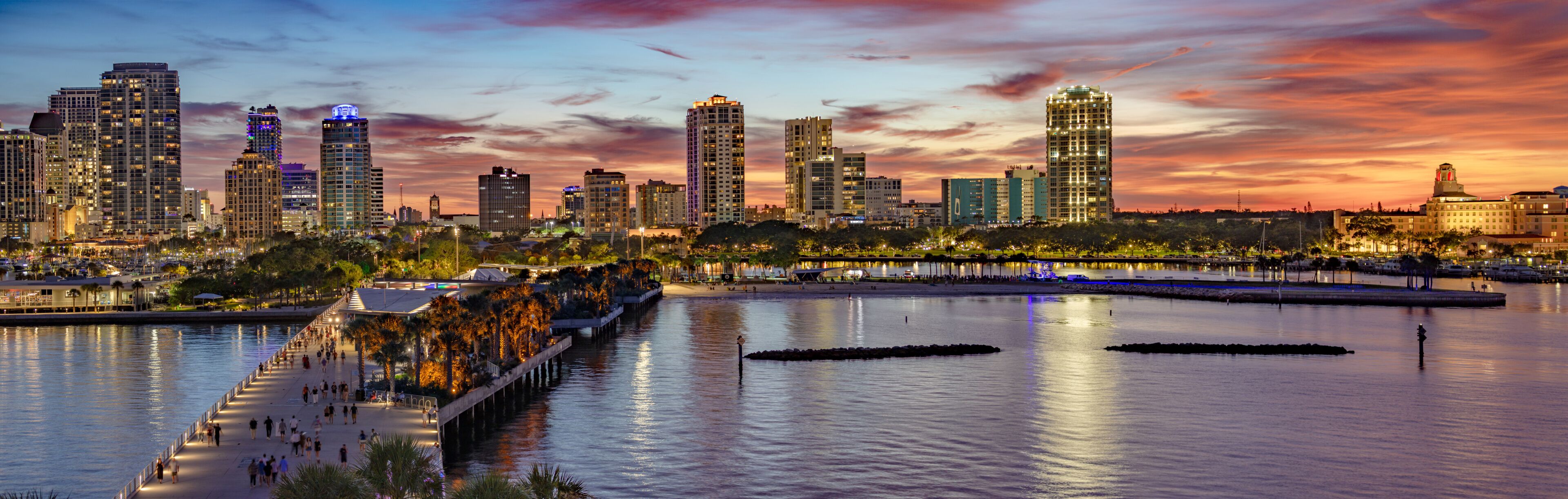 A waterfront cityscape with high-rise buildings and a boardwalk at sunset.