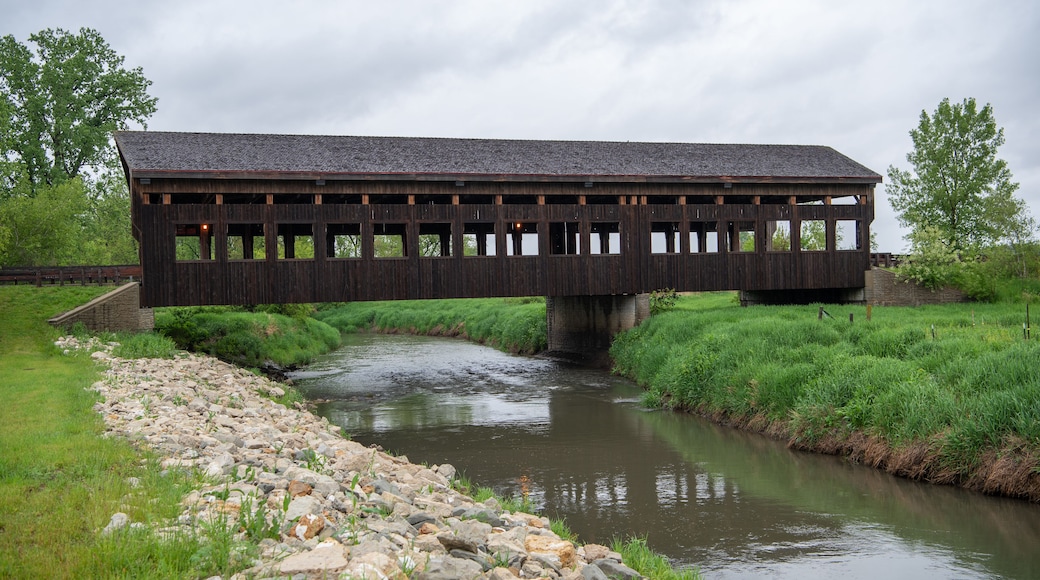 A wooden covered bridge over a calm river with grassy banks and trees in the background.