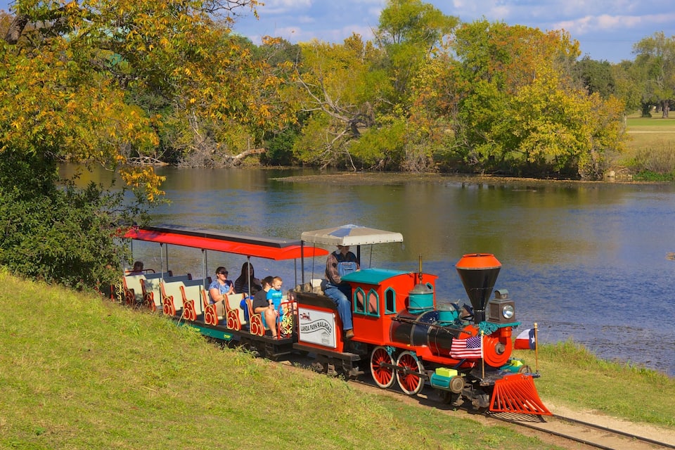 A red and black train with passengers, traveling along a track beside a lake.