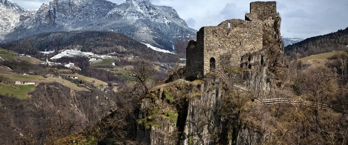 A historic stone castle atop a cliff with snow-capped mountains in the background.