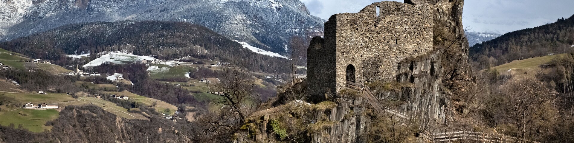 A historic stone castle atop a cliff with snow-capped mountains in the background.