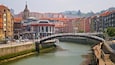 A historic stone bridge over a river, surrounded by colorful buildings and a prominent church with a dome.