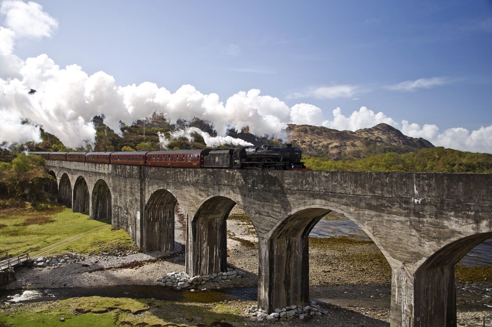 A steam train crossing a stone bridge over a river, with mountains in the background.