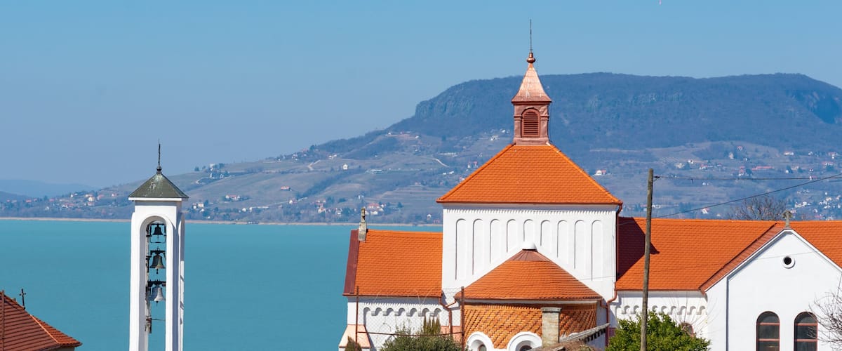 A church with a bell tower and a building with a red roof. In the background, there is a body of water and mountains.
