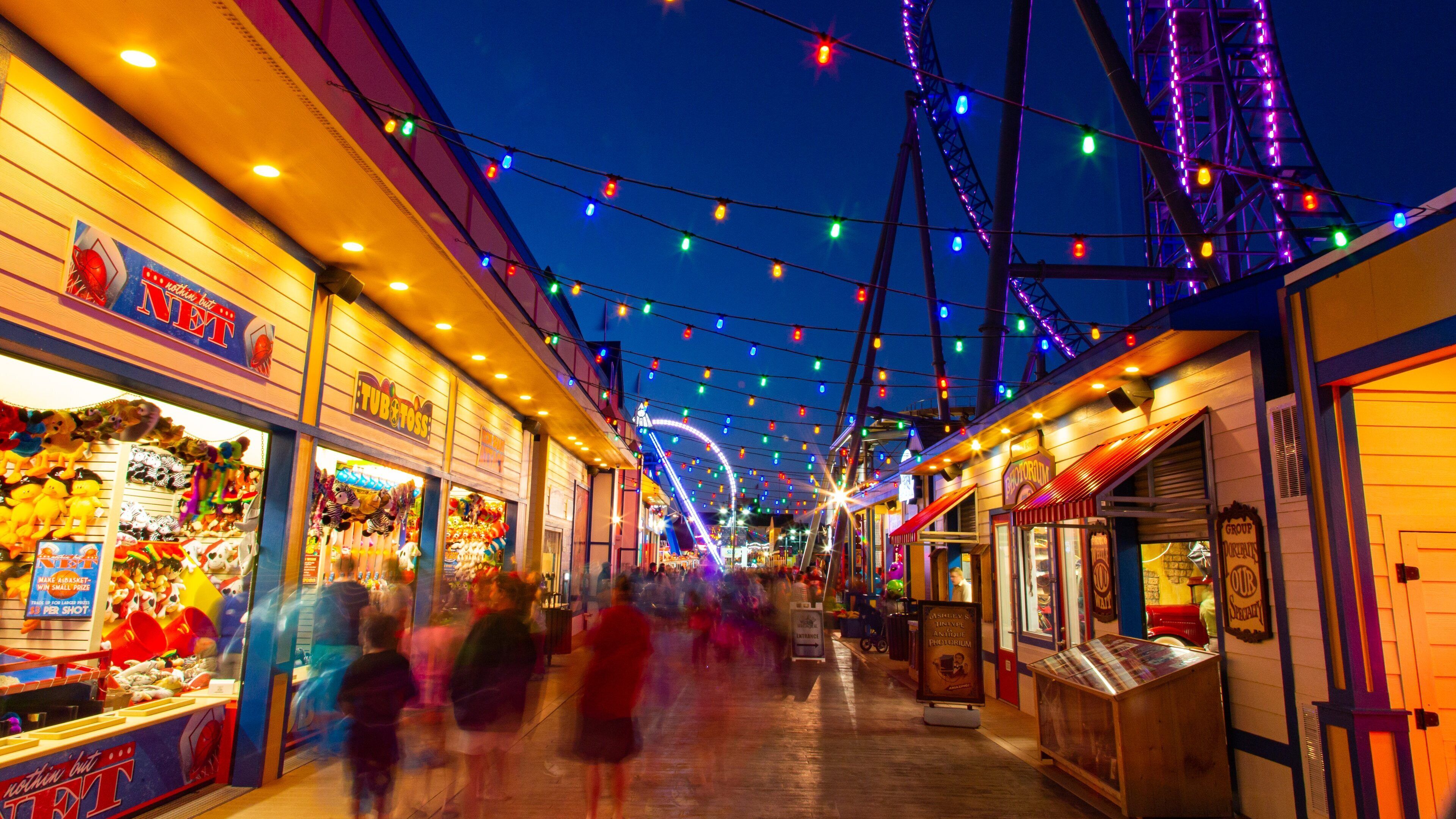 Una calle bien iluminada con luces de colores, tiendas y una noria al fondo.