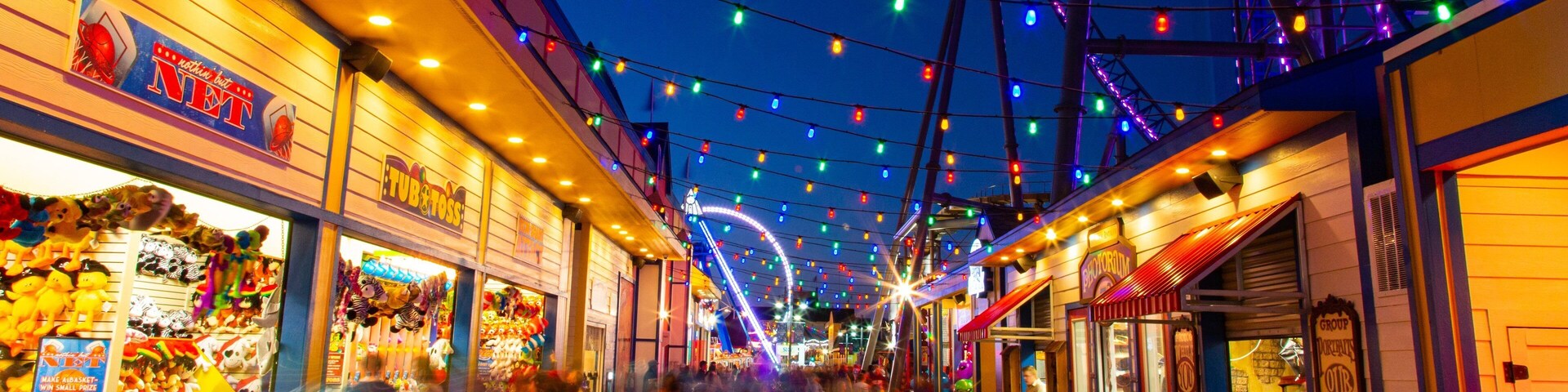 Una calle bien iluminada con luces de colores, tiendas y una noria al fondo.