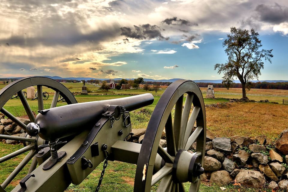 A cannon in a grassy field with a stone wall, a tree, and a cloudy sky.