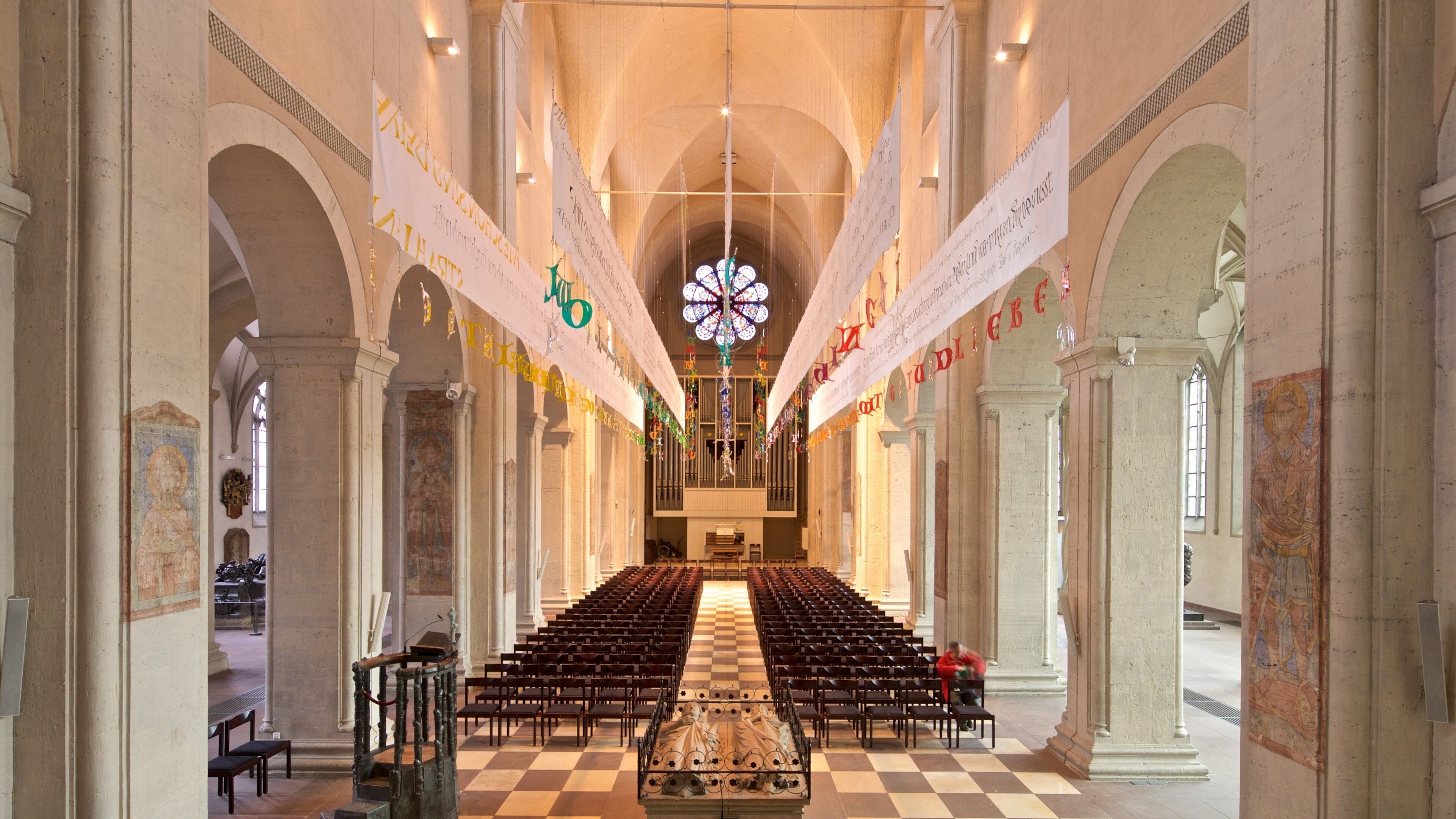 A church interior with rows of pews, a large stained glass window, and banners hanging from the ceiling.