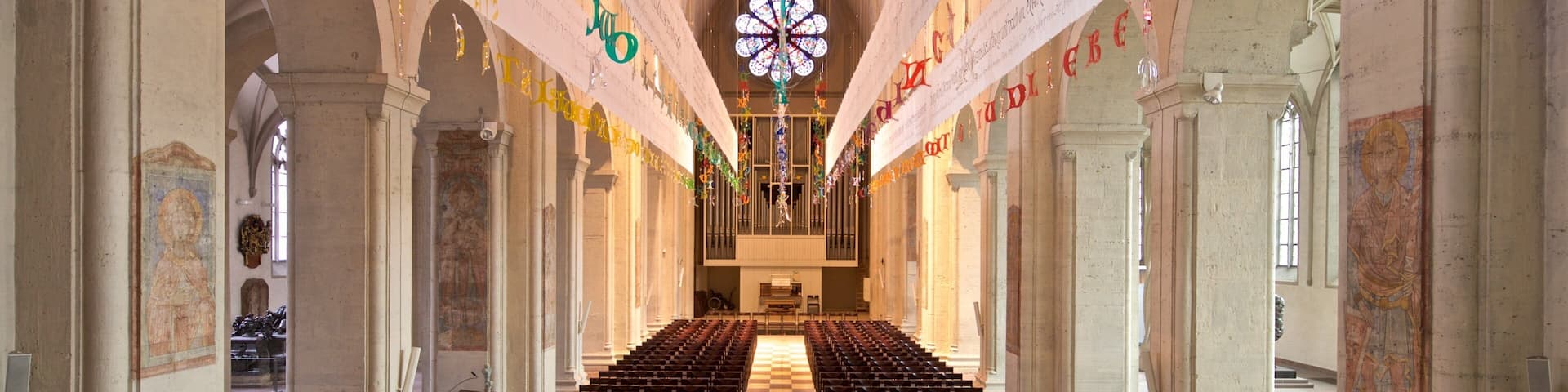 A church interior with rows of pews, a large stained glass window, and banners hanging from the ceiling.
