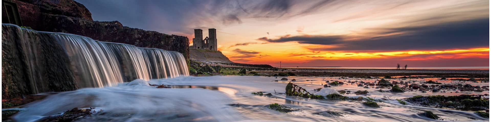 A waterfall, rocky shore, and a castle at sunset.