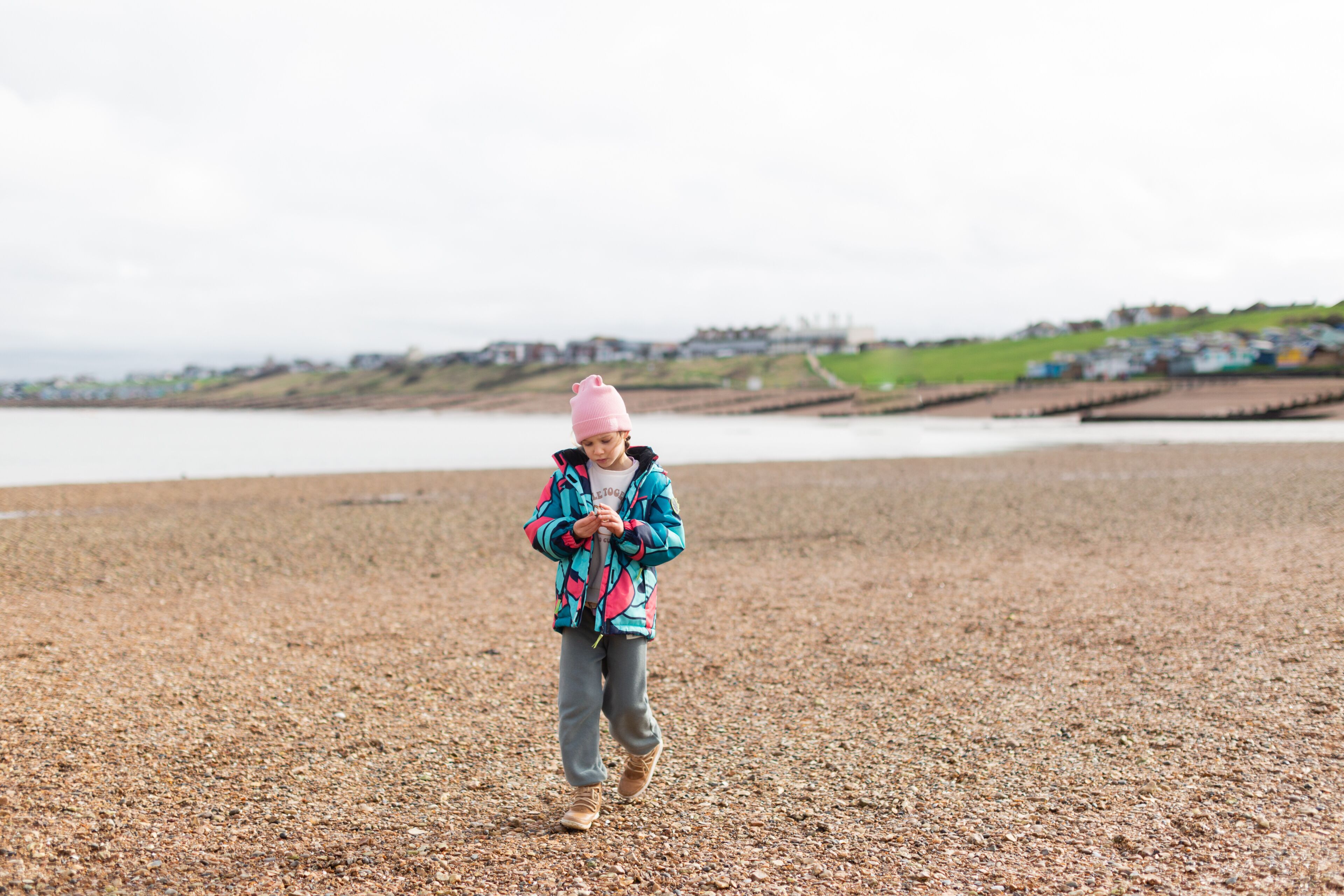 A young girl, wearing a colorful jacket and pink beanie, enjoys a day at the pebbled beach of Whitstable, UK, examining her gathered shells