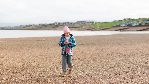 A young girl, wearing a colorful jacket and pink beanie, enjoys a day at the pebbled beach of Whitstable, UK, examining her gathered shells