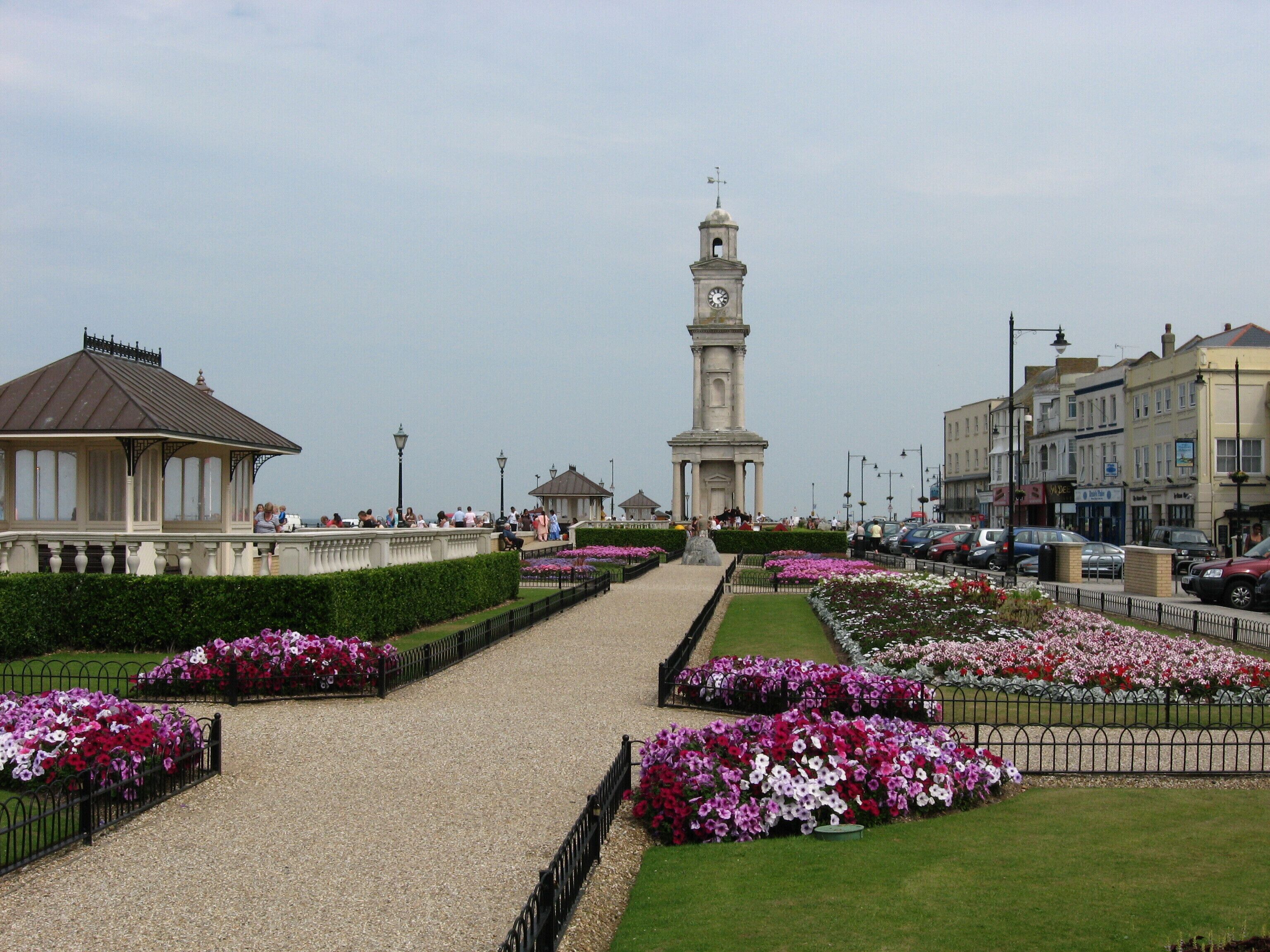A promenade with a central clock tower, flower beds, and surrounding buildings.