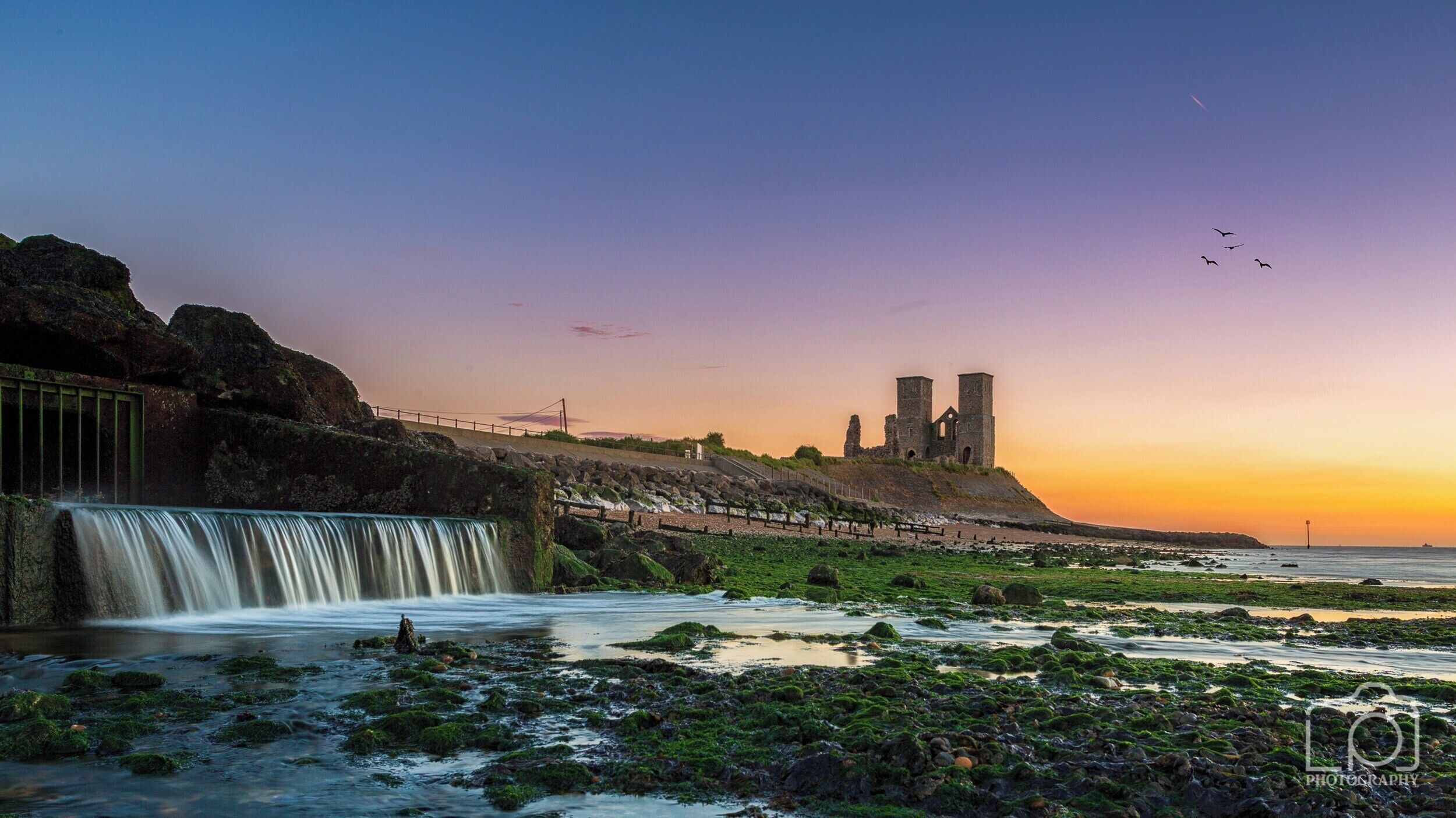 A coastal landscape with a waterfall, rocky shore, and historic ruins.