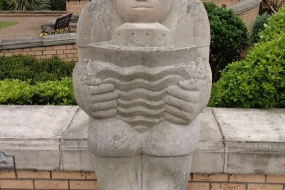 A stone sculpture of a figure holding a shield in a garden with brick walls and greenery.