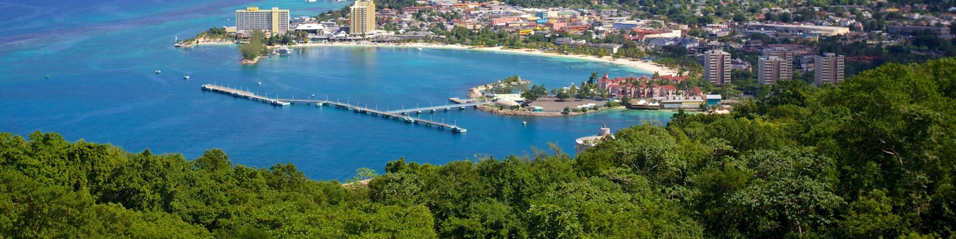 A coastal city with a pier extending into turquoise waters, surrounded by dense green hills and a clear sky.