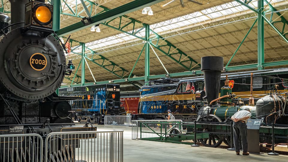 Vintage steam locomotives at a train station with a green metal roof and a sign reading "Please do not climb on the equipment."