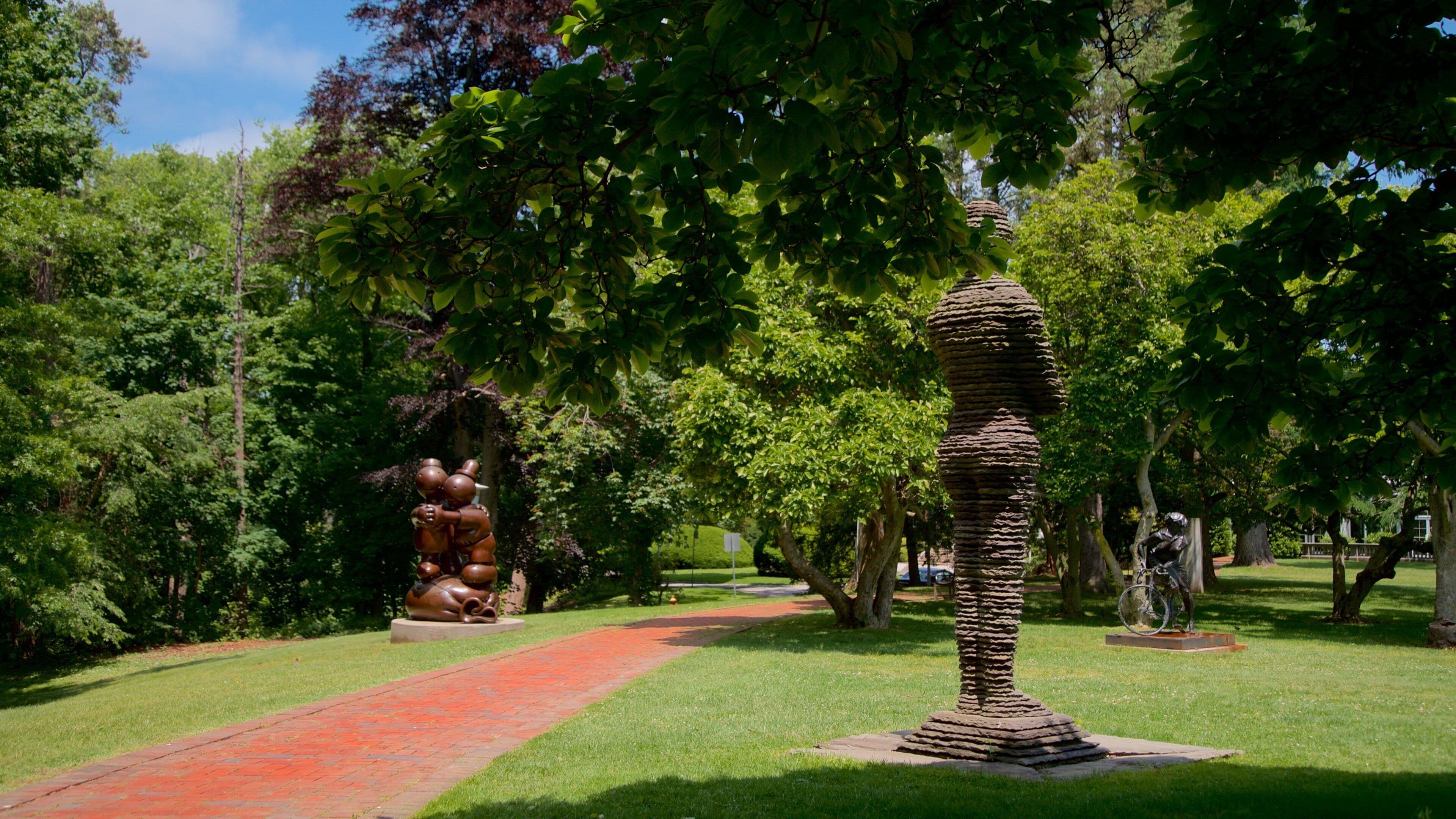 A park with a winding red brick path, a large tree sculpture, and a stone column sculpture.