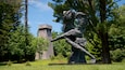 A sculpture of a figure in a dynamic pose, a tower in the background, and a lush green park.