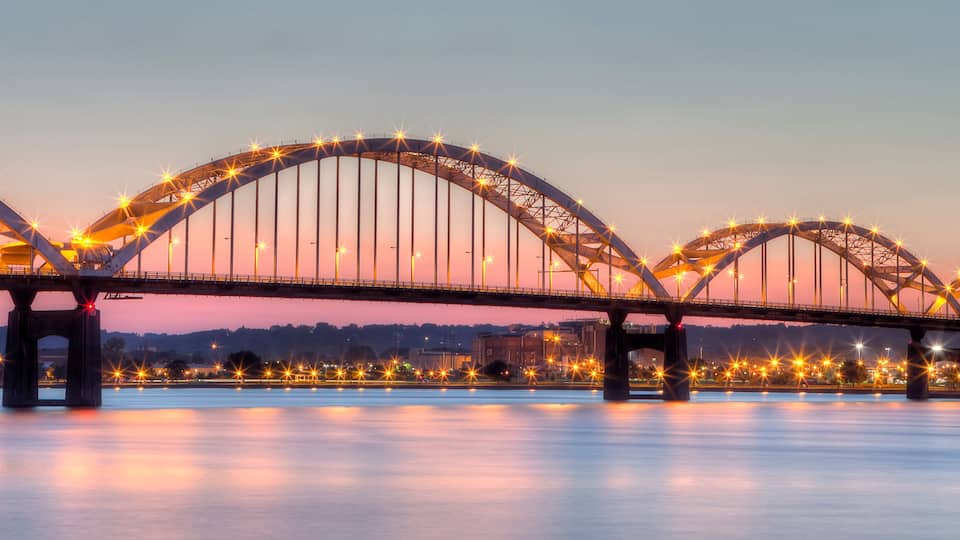 A large illuminated bridge with multiple arches over a calm river at dusk.