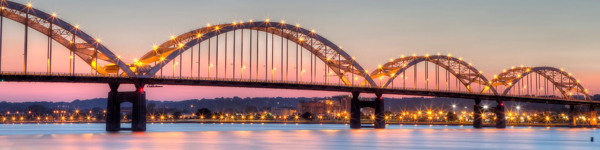 A large illuminated bridge with multiple arches over a calm river at dusk.