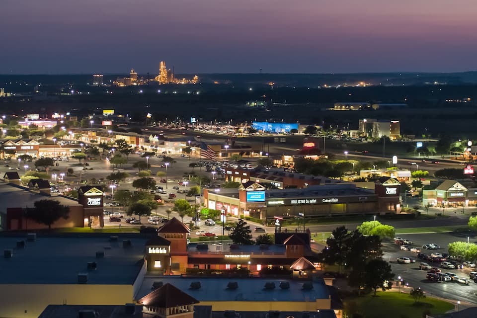 A cityscape at dusk with illuminated buildings, a prominent shopping center, and a distant tower.