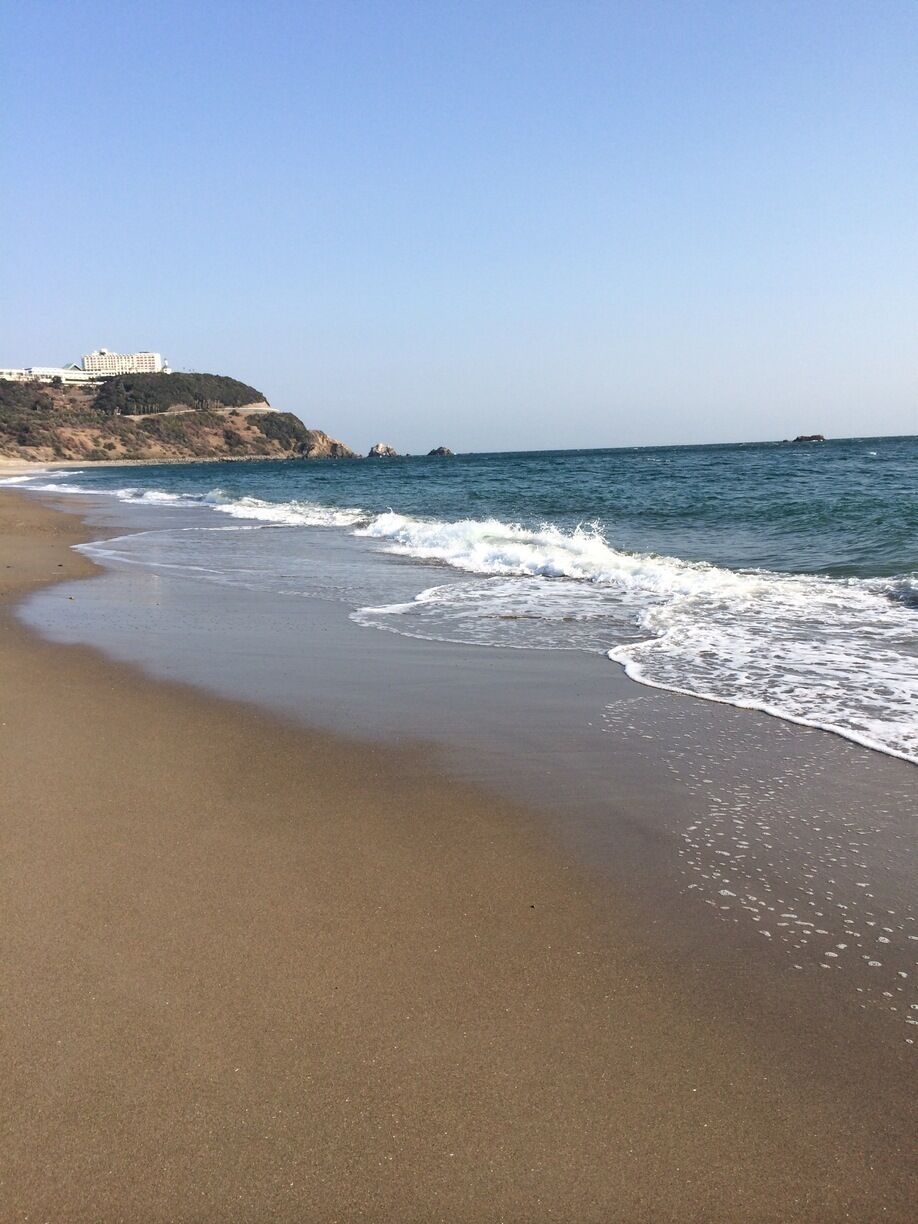 A sandy beach with gentle waves, a clear sky, and a building on a cliff in the background.
