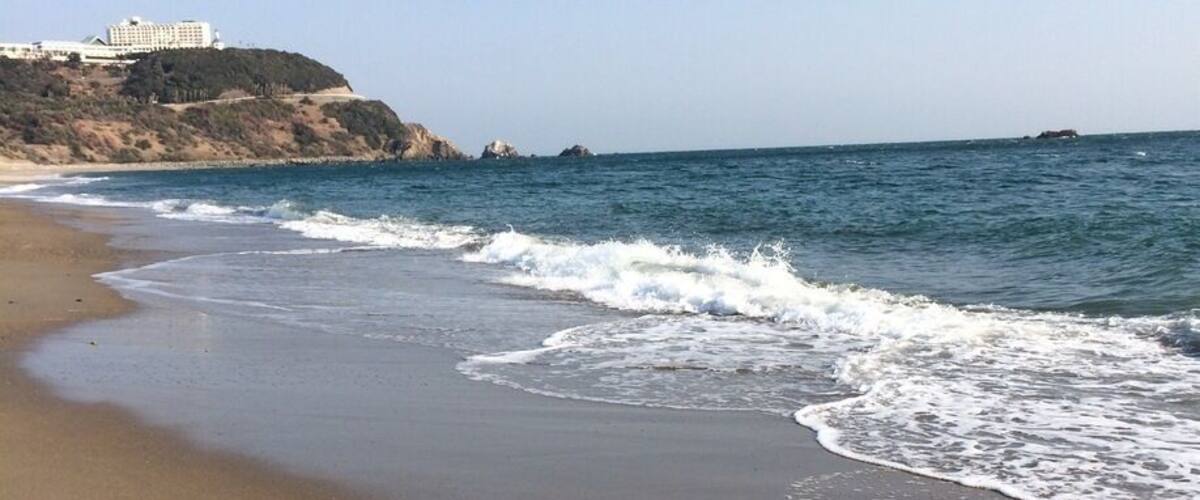 A sandy beach with gentle waves, a clear sky, and a building on a cliff in the background.