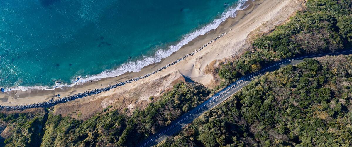An aerial view of a coastal road with a beach and dense forest.