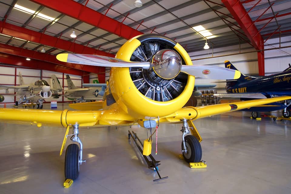 Un avió militar antic de color groc amb unèl·lix, exposat en un cobert amb altres avions.