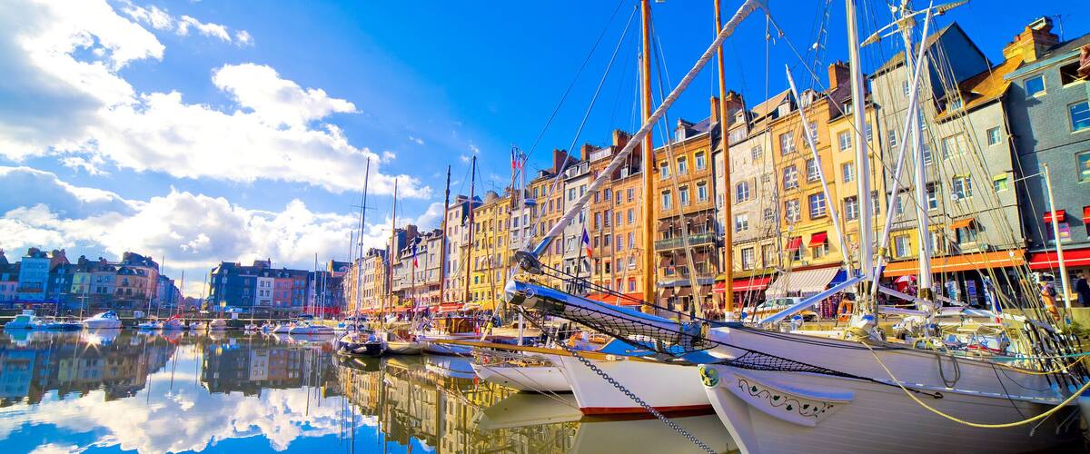 A harbor with multiple sailboats docked, buildings along the waterfront, and a clear blue sky with clouds.
