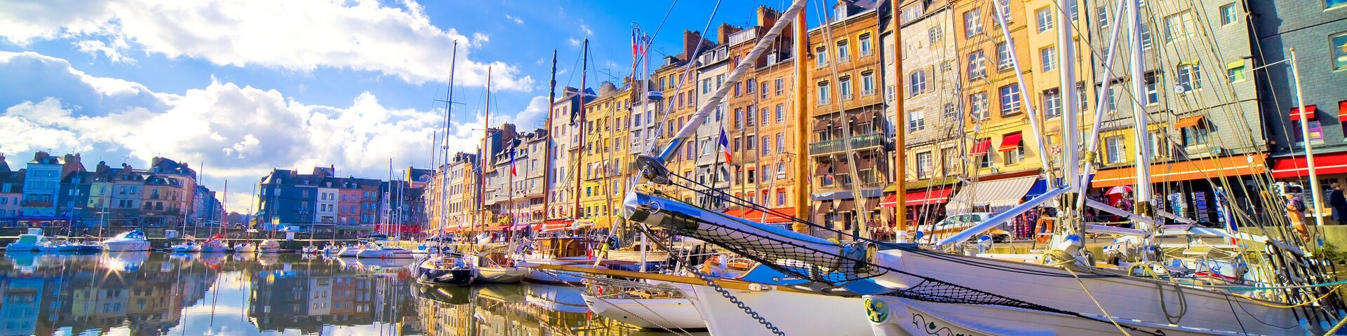 A harbor with multiple sailboats docked, buildings along the waterfront, and a clear blue sky with clouds.