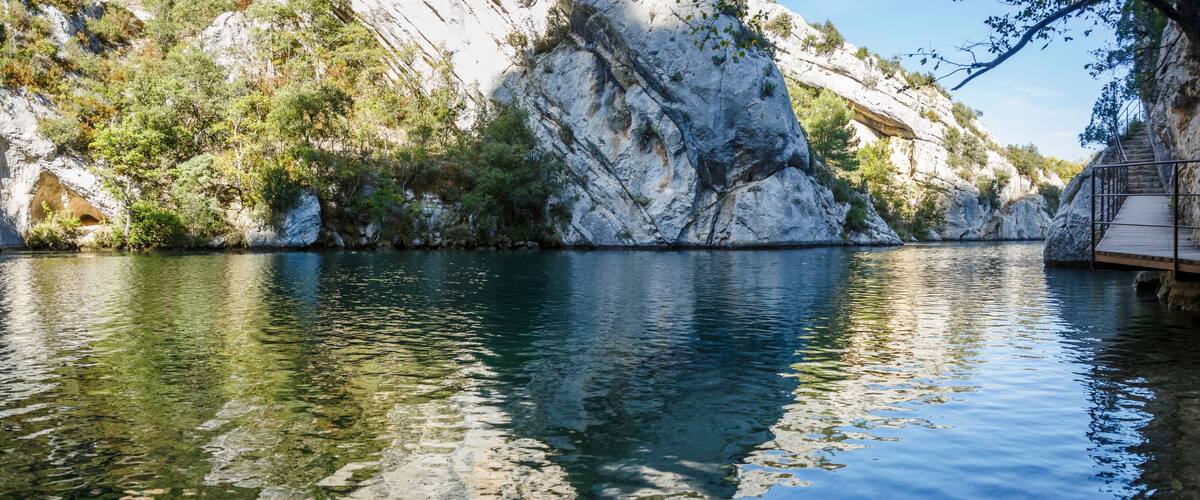 A clear river with rocky cliffs and a wooden walkway.