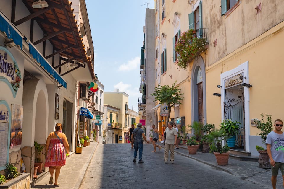 Una stretta via fiancheggiata da edifici colorati, vasi di piante e aree con tavoli all'aperto.