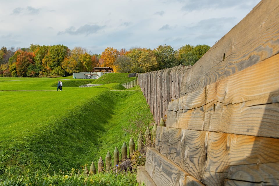 A grassy field with a wooden fence, trees with autumn foliage, and a building in the background.