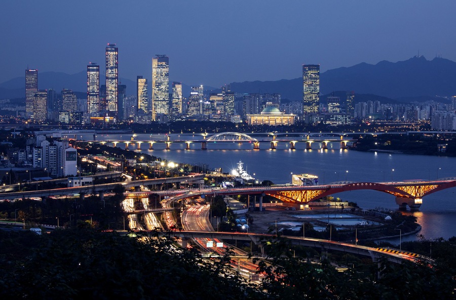 A city skyline at dusk with illuminated buildings, a river, and multiple bridges.