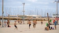 A beach volleyball game with players, a volleyball net, and a backdrop of boats.